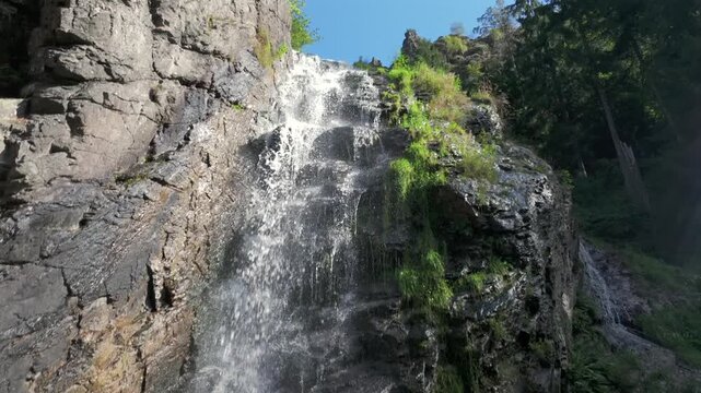 Captivating slow-motion aerial footage of a drone descending along a natural waterfall. The camera smoothly moves downwards, following the path of pure water cascading over a steep rocky cliff in a lu