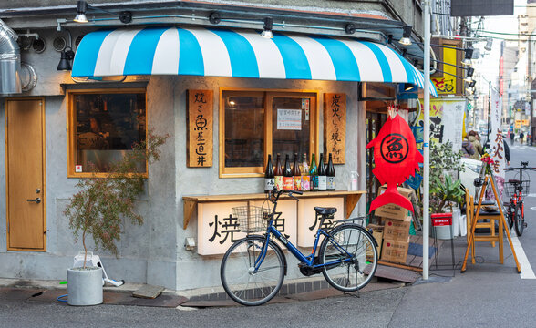 A small restaurant shows a blue and white striped awning with various bottles displayed inside. A bicycle is parked outside next to the entrance on a busy street in the evening