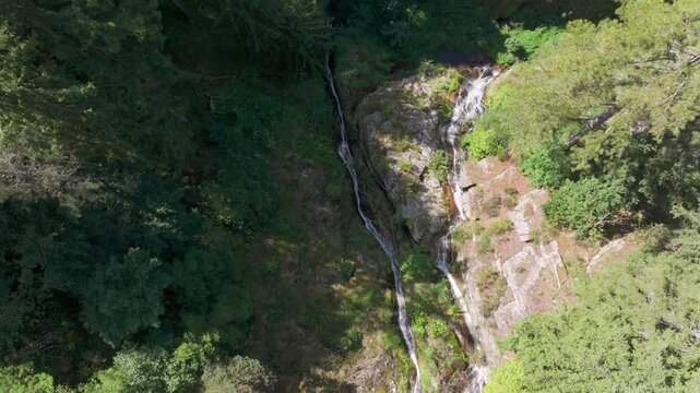 Captivating real-time aerial footage of a drone performing a smooth circular orbit shot around a beautiful wild waterfall. The camera moves dynamically, revealing the waterfall's context within a stee
