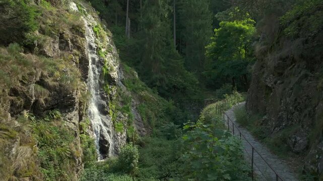 Smooth aerial drone footage moving forward along an old stone walking path with a metal railing, leading towards a beautiful natural waterfall. The fresh water cascades down a rugged rocky cliff insid