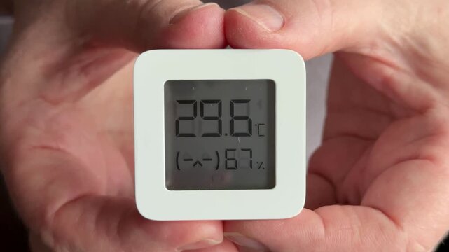 Close-up of woman's hands holding digital sensor displaying temperature and humidity, careful hands tracing display, wrinkled skin.