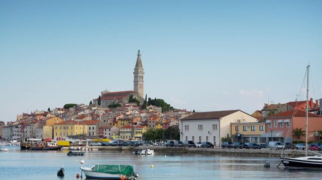 Rovinj, Croatia - August 16, 2025: A tall, historic bell tower rises above the densely packed, terracotta-roofed buildings of Rovinj, overlooking a calm harbor with small boats and parked cars.