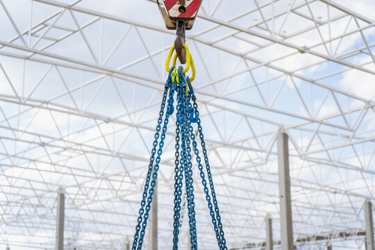Heavy rigging chains attached to crane hook inside industrial building frame with precast columns and roof trusses for construction lifting tasks