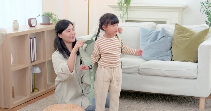 Asian single mother helps preschool daughter prepare backpack in living room before school, guiding routine with care, encouragement, independence training, secure family connection, gentle parenting.