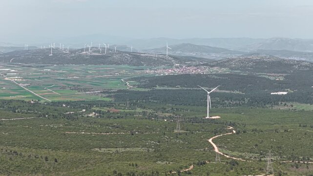 Single wind turbine spinning its blades to generate clean electricity, standing in a vast green landscape with a wind farm and a small village visible in the distant hazy mountains