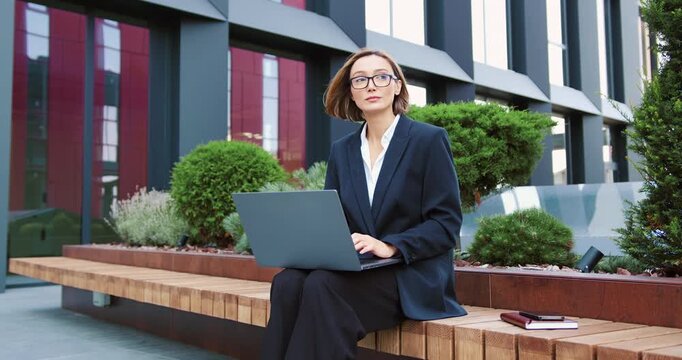 Female entrepreneur in a formal suit sitting on a bench outdoors, typing on a laptop and enjoying remote work in an urban environment