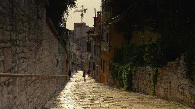 Pula, Croatia - August 17, 2025: A narrow, sun-drenched cobblestone street in Pula winds between ancient stone walls and historic buildings, with people walking toward the warm evening light.