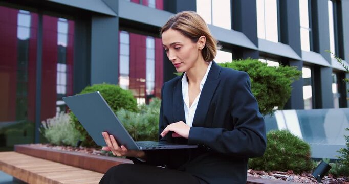 Smiling Caucasian businesswoman working on a laptop outdoors on a bench near an office building, embracing a modern remote work lifestyle.