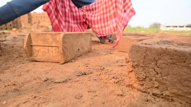 A person manually shaping bricks in an open field. Using a rectangular wooden mold and raw, reddish-brown clay, the worker skillfully forms each brick on the dusty ground. Slow motion video.