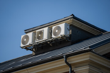 Three rooftop air conditioning condenser units mounted on metal brackets above standing seam roof with gutter and classic building cornice detail © K