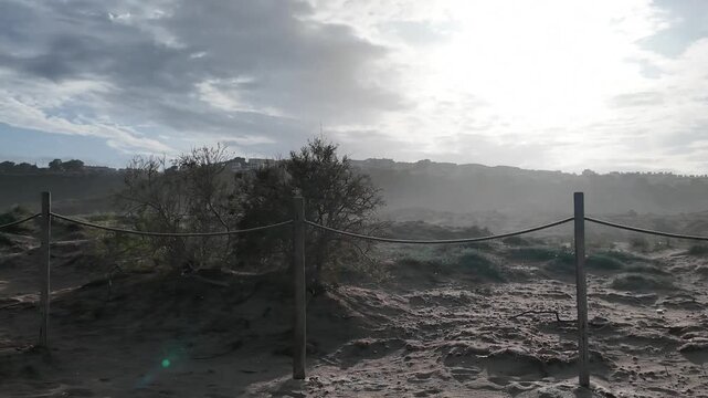 Scenic view of a sandy beach protected by a rustic wooden and rope fence. A beautiful morning landscape showing coastal dunes and distant seaside town houses under a cloudy sky