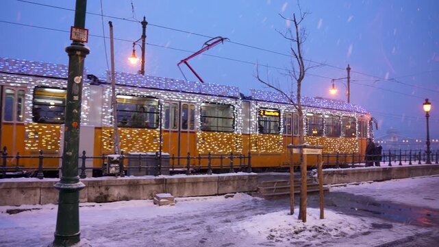 Budapest, Hungary - January 6, 2026: A yellow tram covered in sparkling white holiday lights travels through the snow along the Danube embankment in Budapest during a cold winter twilight.