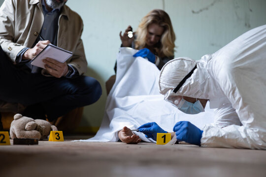 Forensic worker examining a covered body at a crime scene with detectives taking notes and collecting evidence