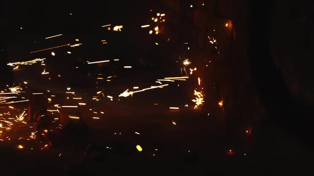 Intense welding sparks scatter across an industrial workshop, illuminating a brick wall, scattered metal components, and a worker's boots during a high-heat metal cutting process.