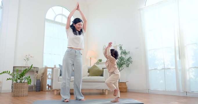 Asian single mother and preschool daughter practice playful yoga at home, balancing together on mat in bright living room, building body control, confidence, bonding, healthy child development