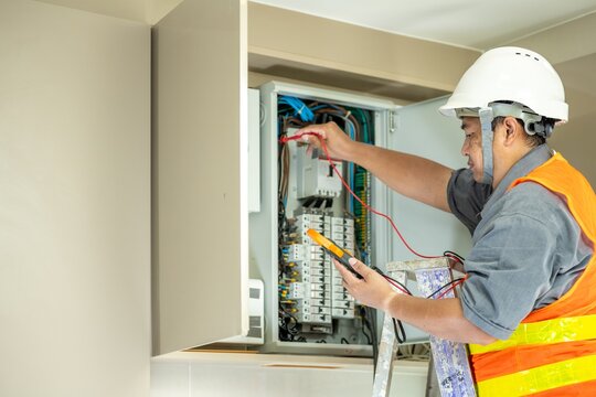 Professional electrician wea safety helmet and reflective vest working on electrical panel, testing wi and electrical connections with multimeter in a commercial building