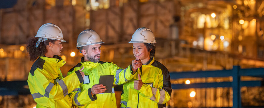 Petroleum engineers team wearing safety uniform and hardhat with laptop monitoring and discussing work at oil refinery site during night operation engineering teamwork in energy industry.