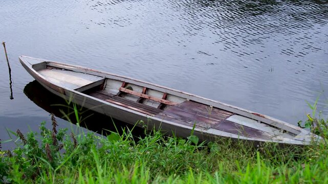Panning shot of an empty traditional wooden fishing boat moored at the grassy bank of Selorejo Reservoir in Malang, Indonesia, representing rural Southeast Asian livelihood and inland fisheries.