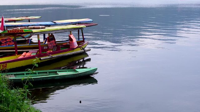 Front view of colorful wooden tourist boats docked at the grassy edge of Selorejo Reservoir, Malang, Indonesia, featuring expansive calm water on the right for ideal copy space and text overlay.