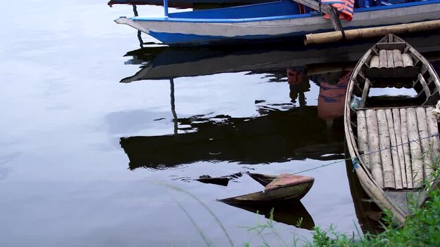 Panning shot of small traditional wooden sampan boats moored at the grassy edge of Selorejo Reservoir, Malang, Indonesia, revealing a broken, sunken boat in the reflective water.