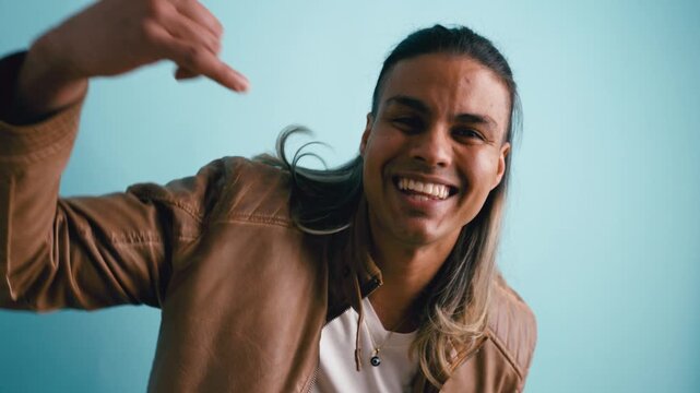 Energetic Latino man with long hair dancing and singing. Joyful guy in a leather jacket rapping and gesturing at the camera in studio
