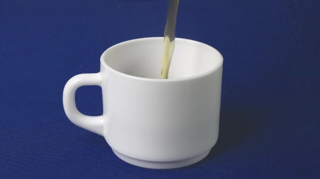A steady stream of dark coffee or tea is poured into a pristine white mug, slowly filling it to the brim on a blue surface
