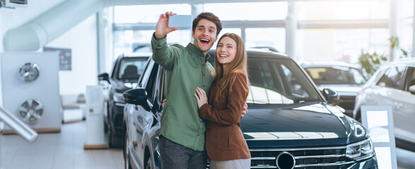 A couple stands close together in a car dealership. They smile and take a selfie. The background shows several cars for sale and bright lighting from large windows. © Prostock-studio