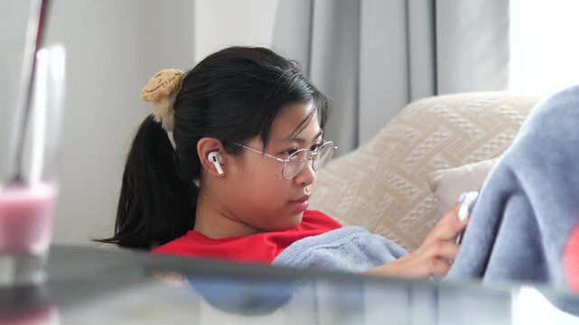 Close-up of a young Asian girl wearing glasses and wireless earbuds, relaxing on a sofa while using a digital tablet at home, digital lifestyle and leisure concept