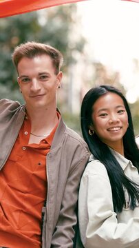 Portrait of a young diverse couple, an asian woman and a caucasian man, smiling at the camera standing back to back under a rainbow lgbt flag. Vertical footage