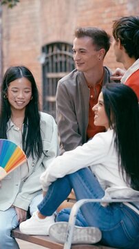 Young group of diverse friends laughing and chatting on a bench. One person is holding a rainbow flag, another a rainbow fan, celebrating pride. Vertical footage
