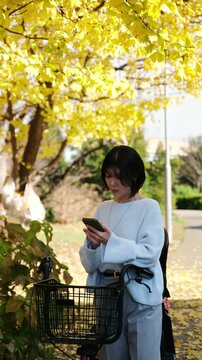 Young asian woman with a bicycle checking her phone under yellow ginkgo trees in autumn