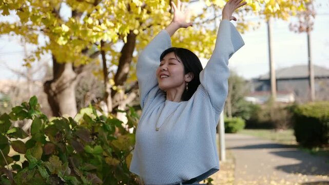 Young Asian woman stretching and enjoying a sunny autumn day in a park with yellow ginkgo trees