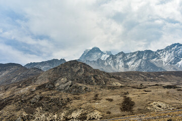 Fototapeta premium Rugged high-altitude landscape in North Sikkim, India, featuring rocky terrain, barren slopes, and distant snow-capped Himalayan peaks under a cloudy sky.