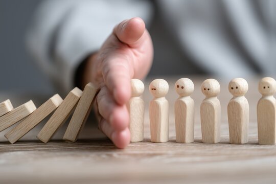 a person's hand is shown in a close up, eye level shot, preventing a chain reaction of light brown wooden dominoes from striking three smiling wooden peg dolls Generative AI