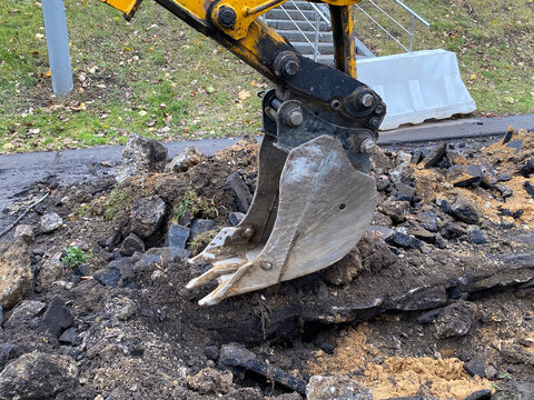 Excavator bucket digging up asphalt and soil during road works. Metal backhoe scoop. Industrial machinery removing old pavement and earth. Hydraulic excavator working on urban infrastructure repair.