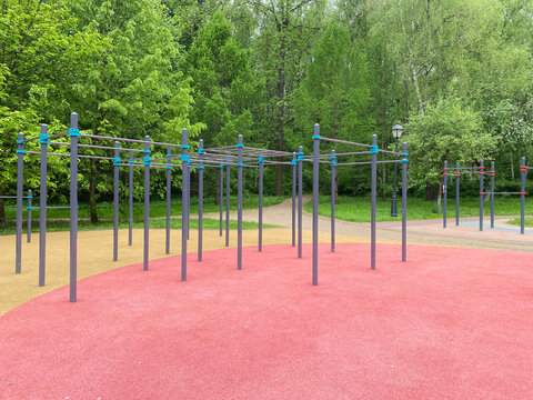 Street workout station featuring metal horizontal bars and monkey bars installed on red and yellow rubber safety flooring. Modern fitness equipment located in green summer park with lush trees.