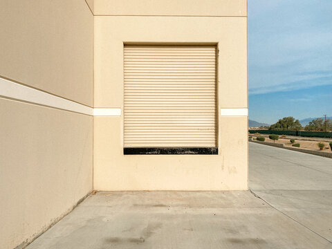 A beige industrial building with a closed roll up door sits on a clean concrete driveway under a blue sky with mountains and trees in the background the scene is empty