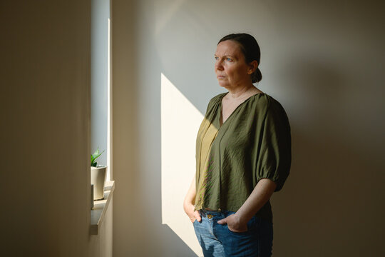 Pensive woman illuminated by soft window light, looking out the window with introspective expression, emotional lifestyle portrait in a calm home interior.