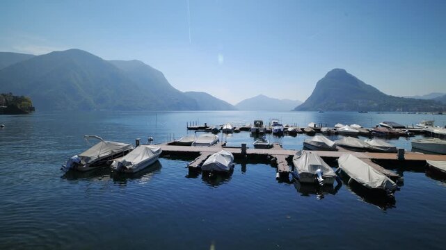 Wide shot of boats at Lake Lugano pier with scenic mountain peaks under a clear blue sky