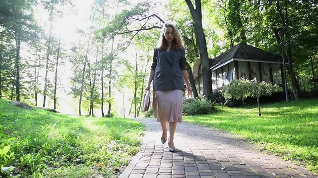 Slow motion shot of a young blonde woman walking in a park when her heel slips and she almost falls, laughing loudly. Bright sunlight and candid reaction create a playful lifestyle moment.
