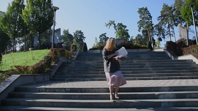 Slow motion shot of a young blonde woman walking down stairs while looking at documents as loose papers fall to the ground. Bright sunlight and greenery create a natural outdoor lifestyle scene.