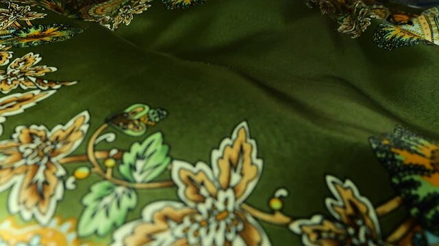 Green silk fabric with floral pattern develops in the wind. Waving textile material creates abstract background with soft folds. Silk surface features traditional ornaments in yellow and brown colors.