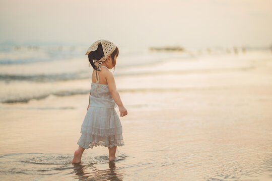 Adorable Asian toddler girl in white boho dress walking on beach at sunset. Cinematic golden hour lifestyle photography of child exploring seaside during summer vacation. Perfect for travel, parenting