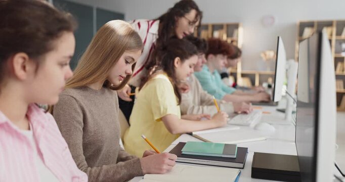Teacher guides students teenagers in classroom computer study. She guides teens as they type and take notes at desktops, building coding and research learning. Guided project teamwork in class.