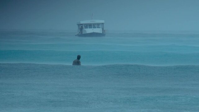 Lonely Surfer Sits In Stormy Ocean, Facing Distant Boat Through Heavy Rain And Mist Moody Teal Tones, Overcast Sky, Soft Swells, Contemplative Pause And Quiet Isolation