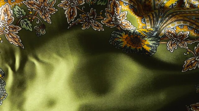 Natural silk fabric with paisley pattern waving in the wind. Green textile material creates abstract waves background. Floral ornaments in yellow and brown colors on smooth surface.