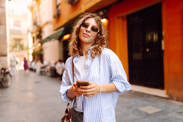 A happy tourist with a smartphone stands on a sunny street. A beautiful woman admires the city's architecture with her phone at sunset. Concept of tourism, recreation, and vacation. © maxbelchenko