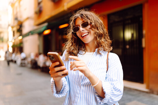 A happy tourist with a smartphone stands on a sunny street. A beautiful woman admires the city's architecture with her phone at sunset. Concept of tourism, recreation, and vacation.