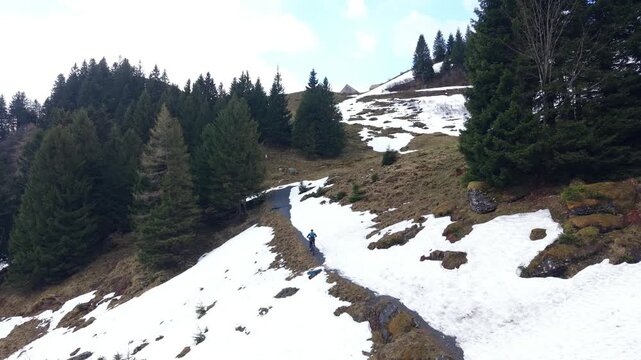 Drone static shot of a cyclist continuing uphill after passing a snowy section, riding away on a partially clear mountain road