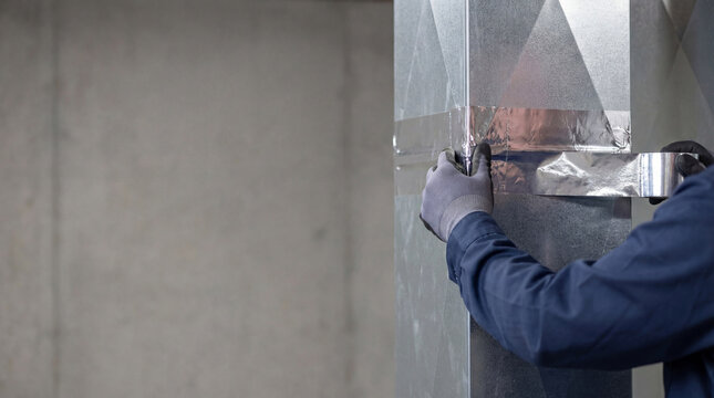 Technician applying silver foil tape to metal air conditioning duct. Professional man installing hvac ventilation system in building. Construction work and climate control maintenance.
